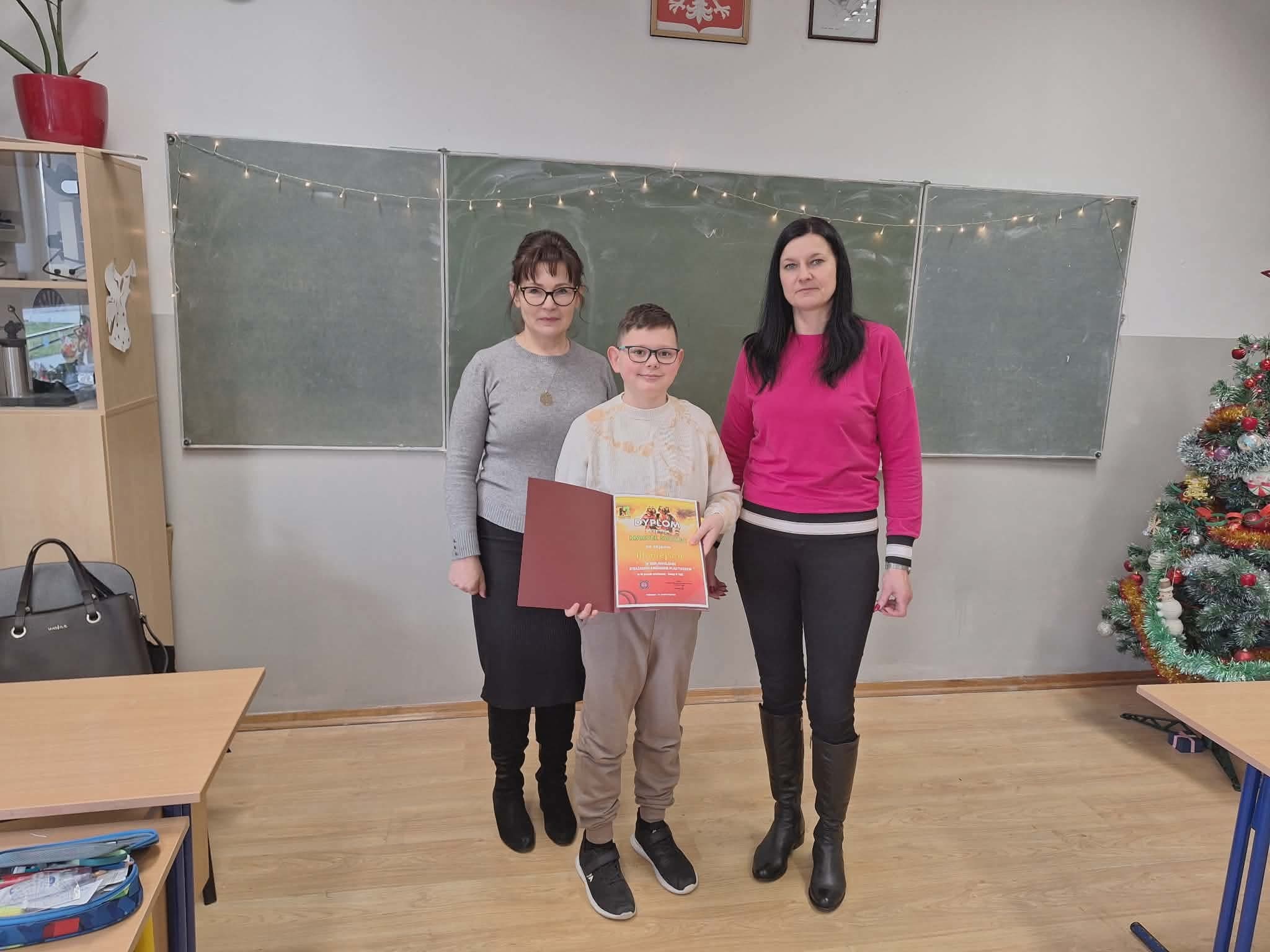 Uczeń z dyplomem konkursowym podczas pamiątkowego zdjęcia z nauczycielkami w sali lekcyjnej.